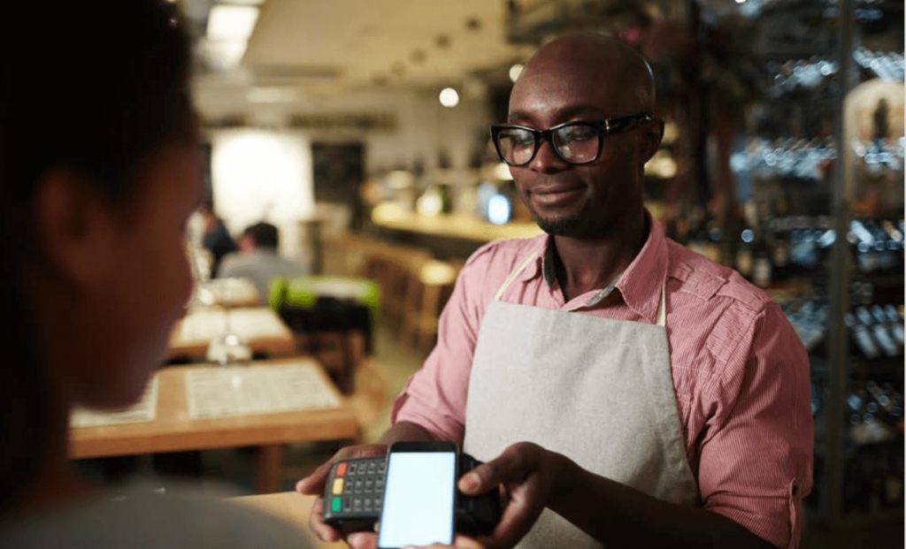 A photo of a restaurant server holding a payment terminal as a customer completes a mobile payment in a warmly lit dining space.