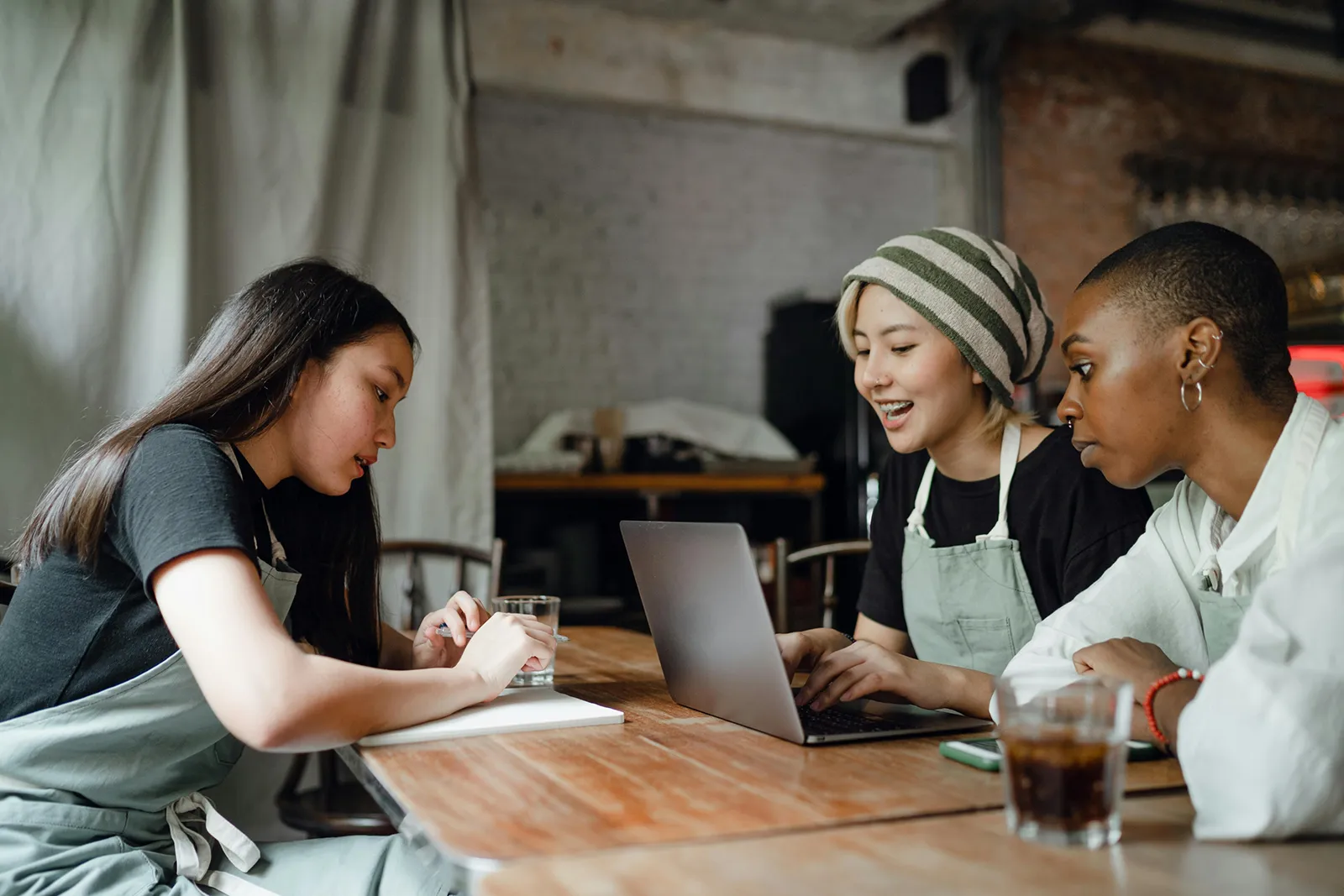 Three café staff members wearing aprons sit at a wooden table with a laptop, notepad, and drinks, collaborating and reviewing information together.