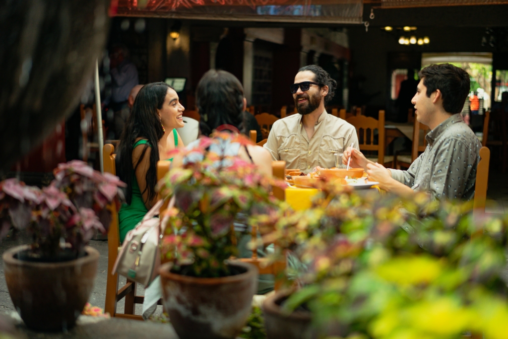photo of friends laughing and talking at an outdoor restaurant table surrounded by plants.