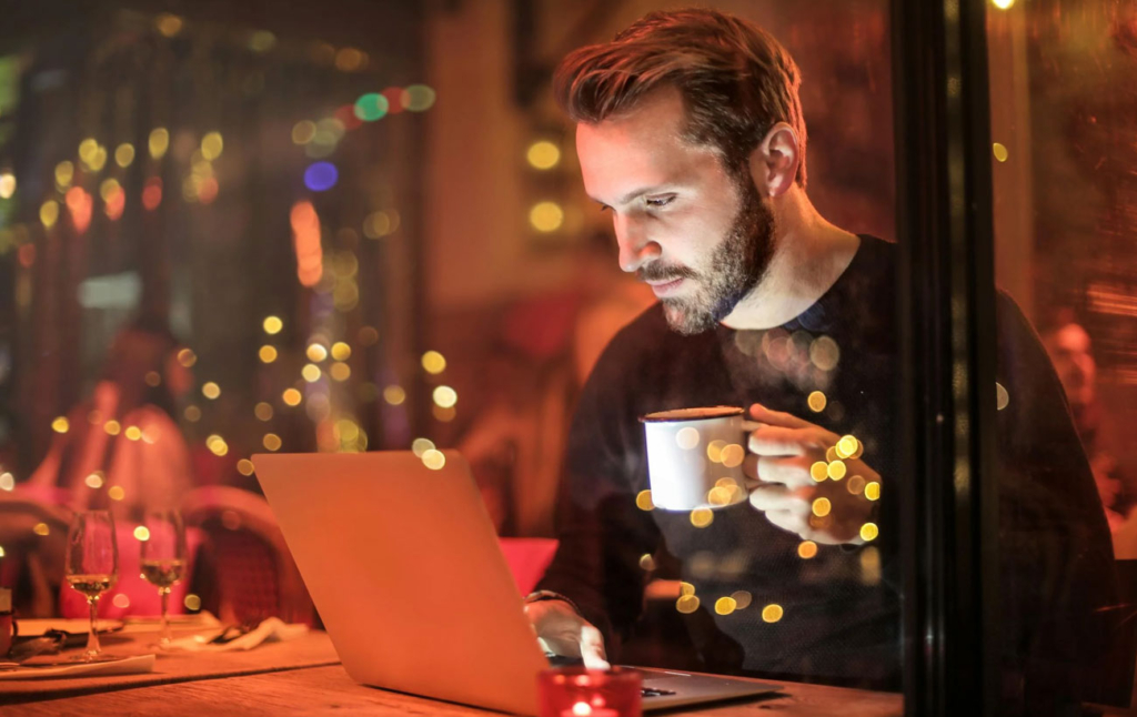 a person working on a laptop and holding a coffee mug inside a warmly lit café.