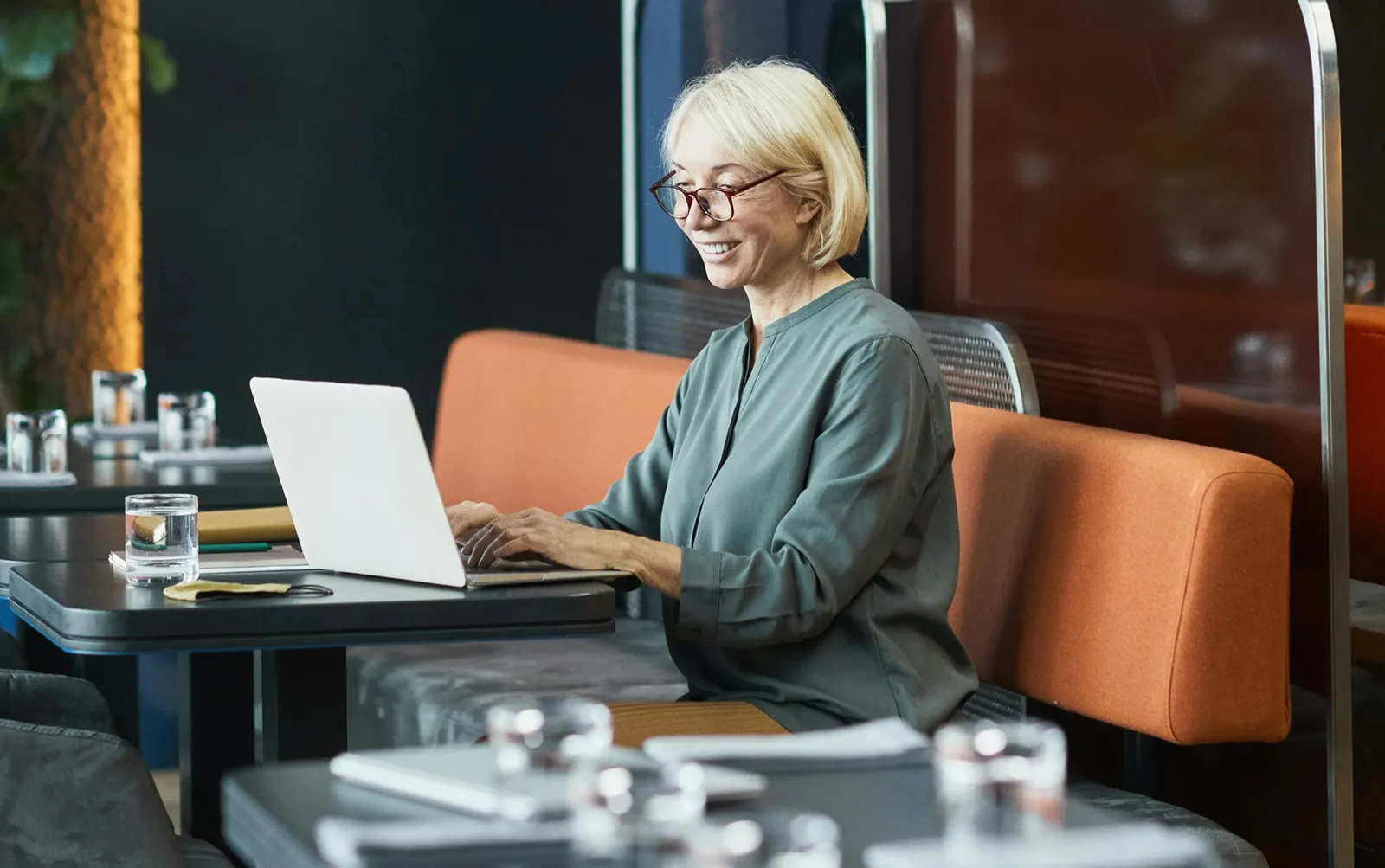A woman with glasses working on a laptop at a café table, smiling as she types.