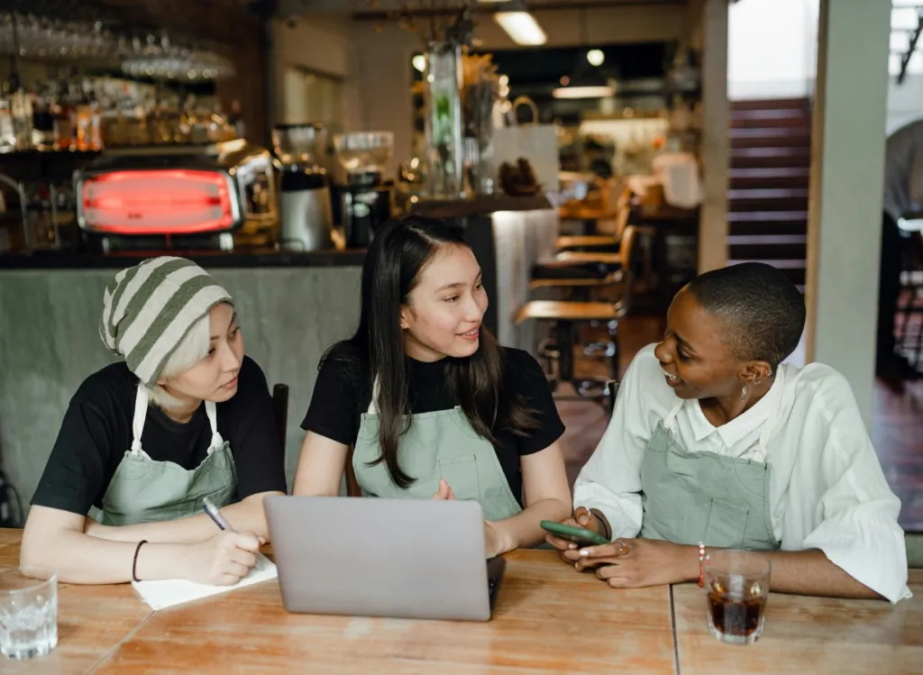Three restaurant staff in aprons sitting at a table with a laptop and phone, smiling and collaborating.