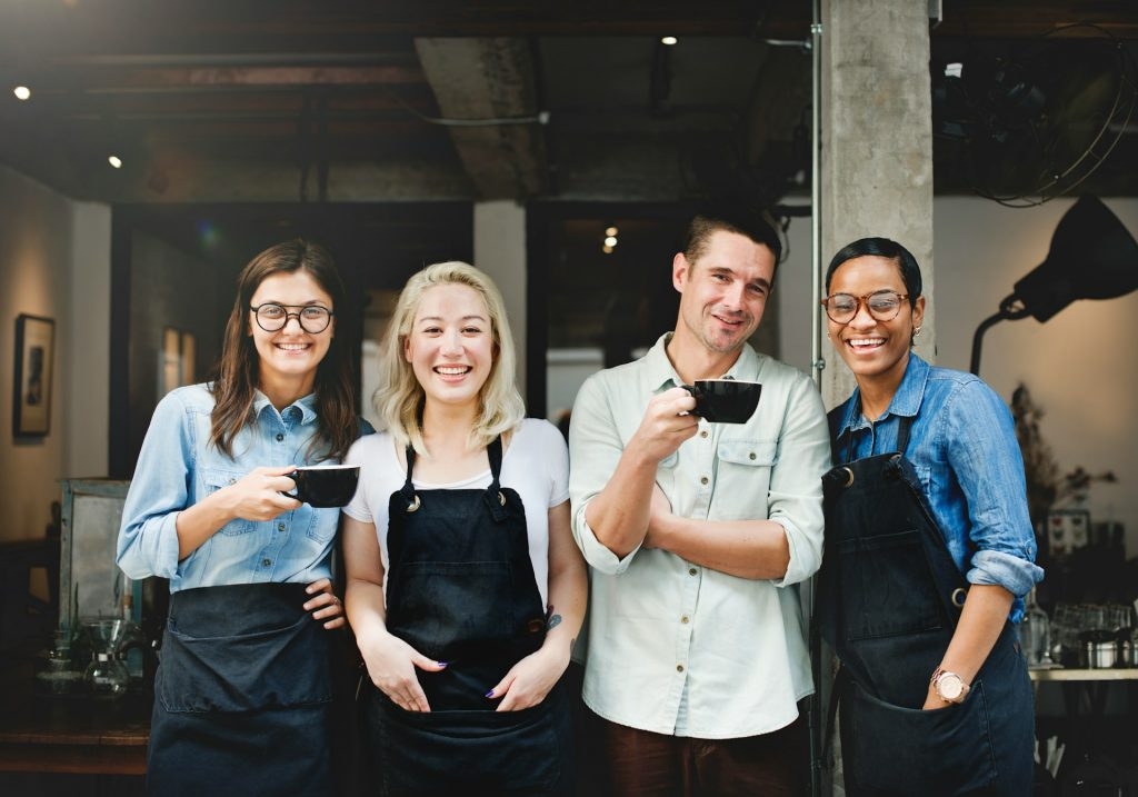 Group of four smiling restaurant staff in aprons standing together with coffee cups.