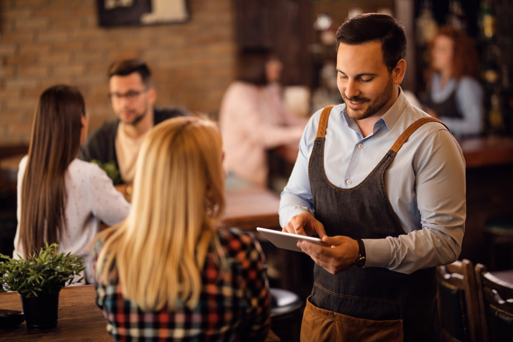 Male server in apron taking an order on a tablet from a guest at a restaurant table.