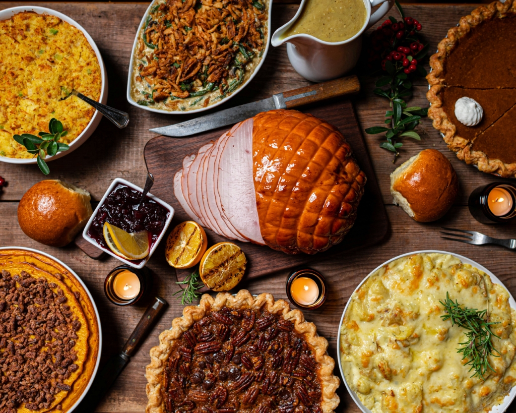 An overhead view of a Thanksgiving feast featuring sliced ham, pies, cranberry sauce, and classic side dishes on a wooden table.