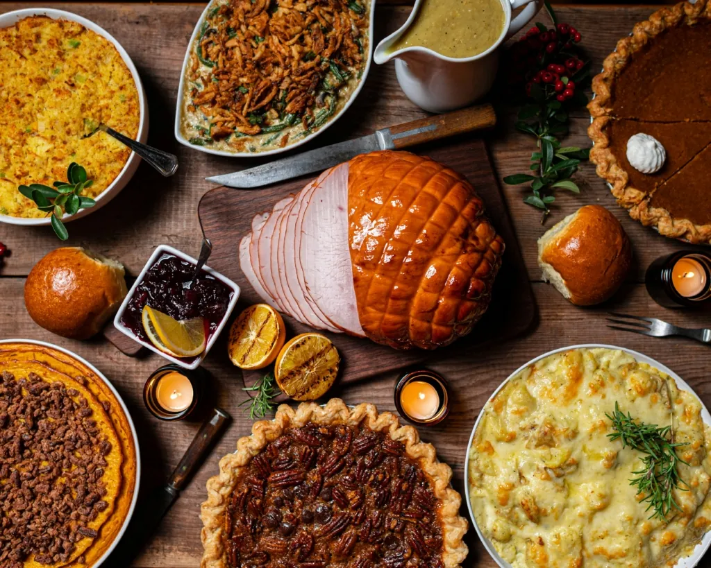 An overhead view of a Thanksgiving feast featuring sliced ham, pies, cranberry sauce, and classic side dishes on a wooden table.