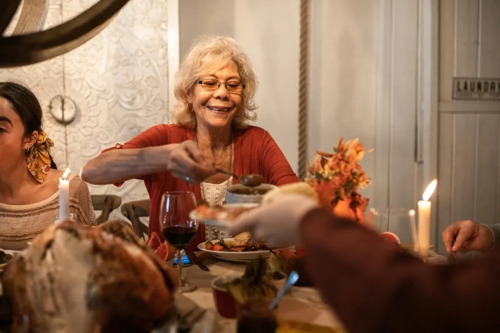 An older woman smiles as she serves food at a cozy Thanksgiving dinner table, surrounded by family and candlelight.