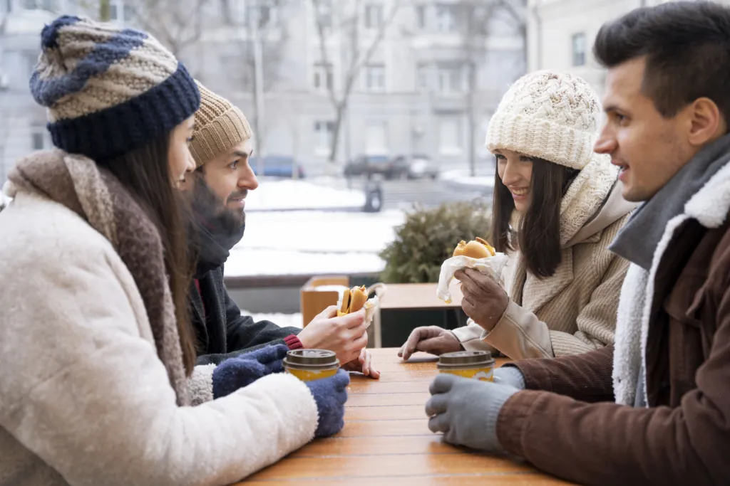 A group of friends in winter coats and beanies enjoy hot drinks and sandwiches together at an outdoor café on a snowy day.