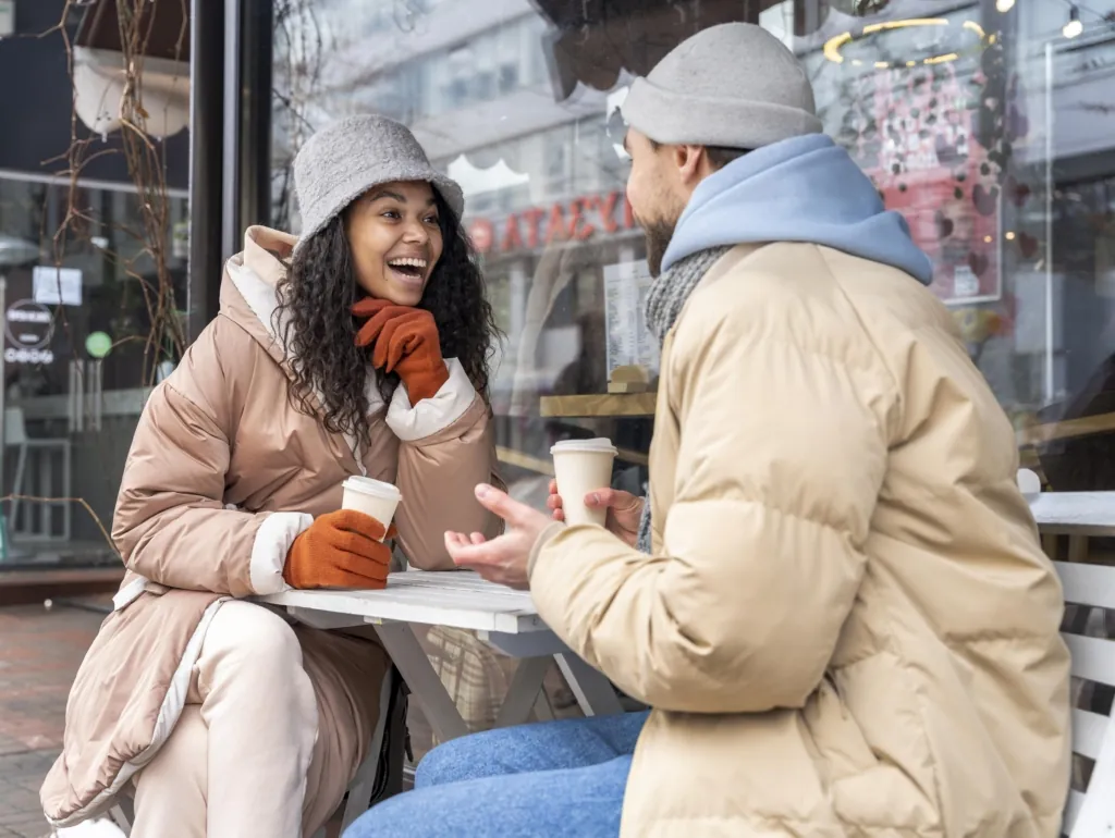 Two friends dressed warmly in winter coats laugh and talk while enjoying coffee outside a café.