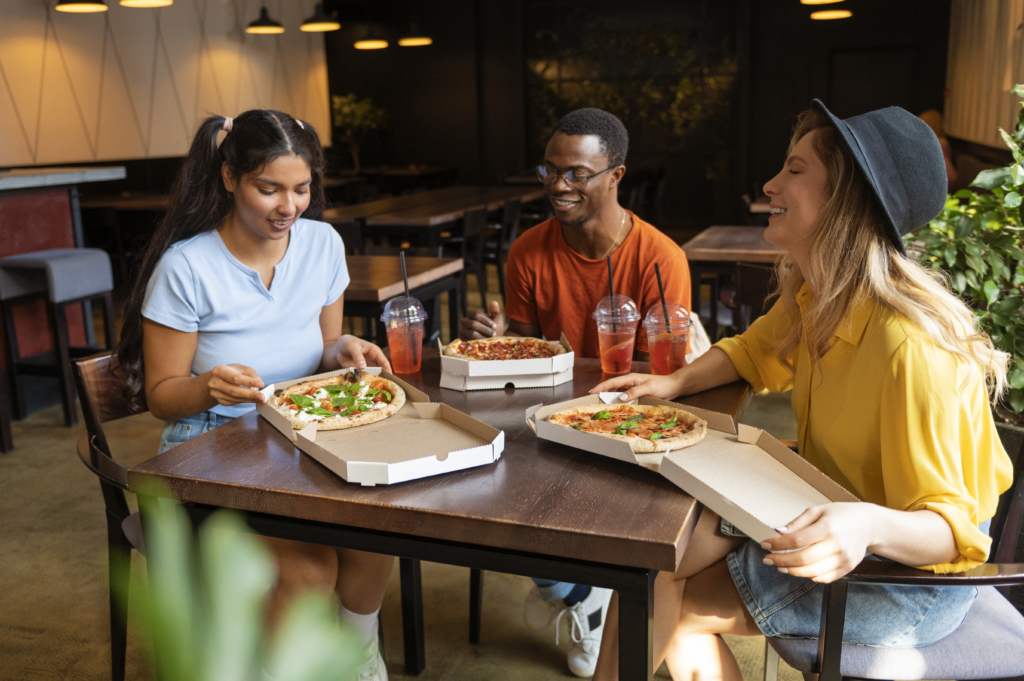 a photo of three friends sitting in a restaurant enjoying pizzas from takeout boxes with iced drinks on the table.