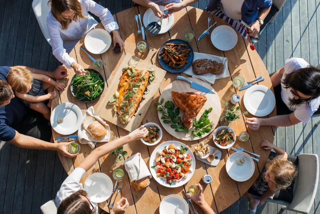 a photo of a group gathered around a large outdoor table sharing a meal with dishes like salmon, roasted vegetables, salad, and bread