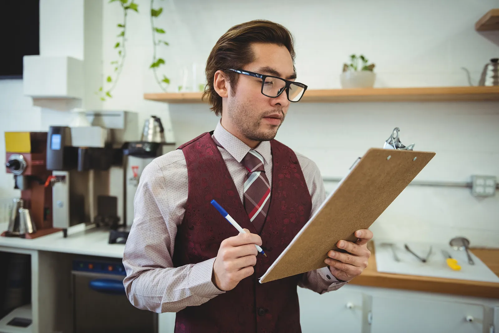 Barista in a vest and tie standing behind a café counter, holding a clipboard and pen while reviewing a checklist, with coffee equipment and shelves in the background.