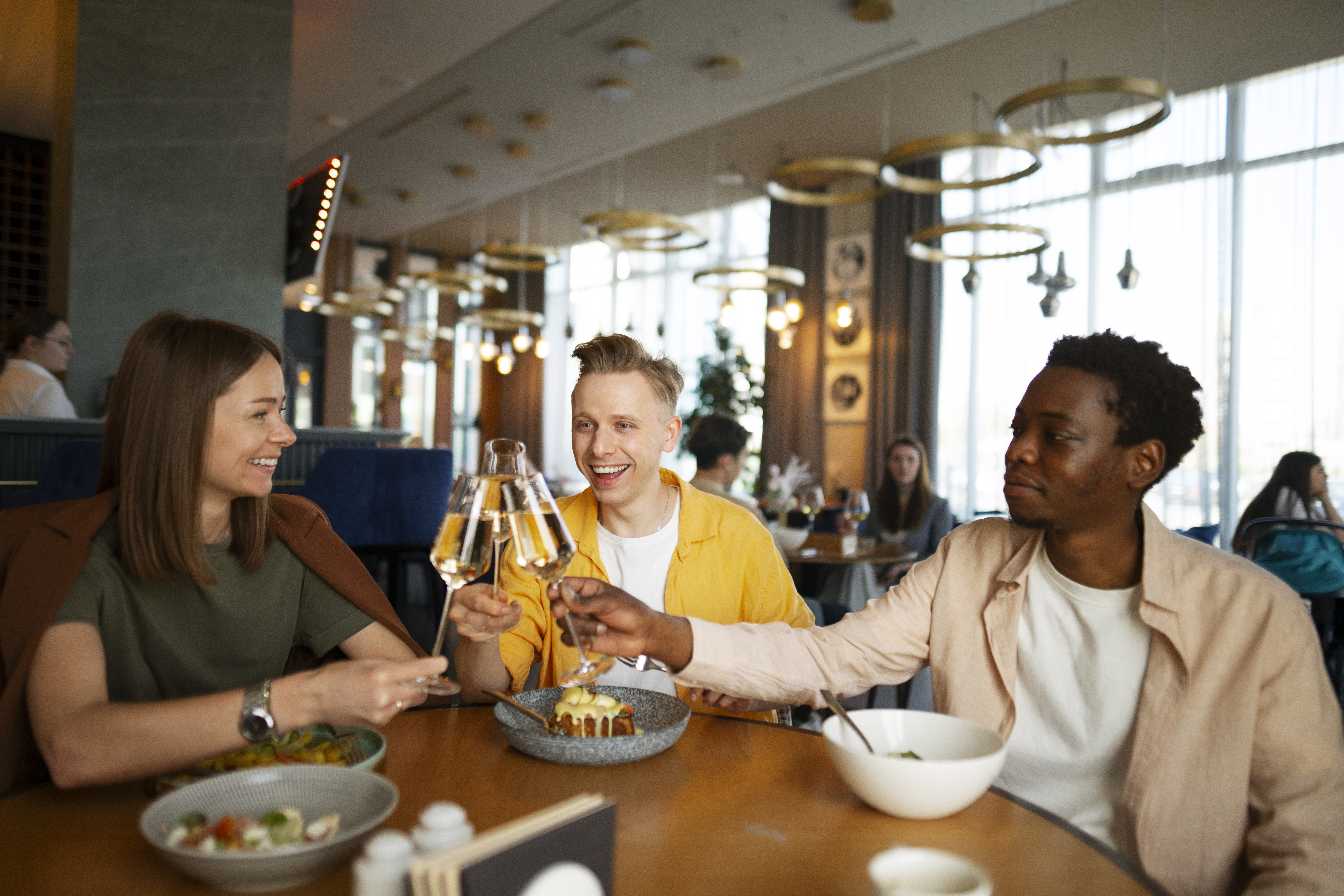 a photo of three friends in a restaurant raising glasses in a toast while enjoying a meal together.