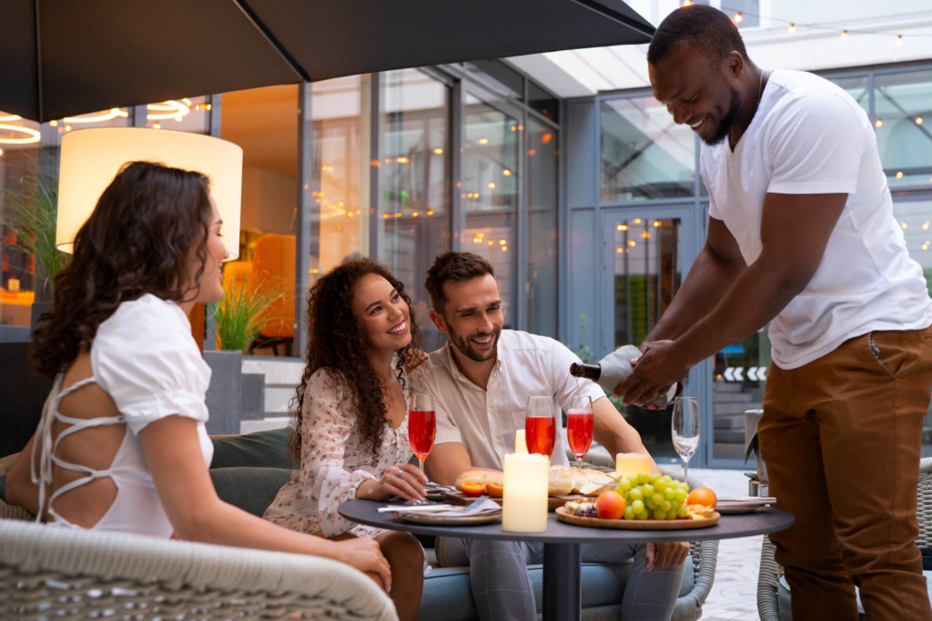 a photo of a server pouring drinks for three guests seated outdoors at a restaurant table with food and candles.