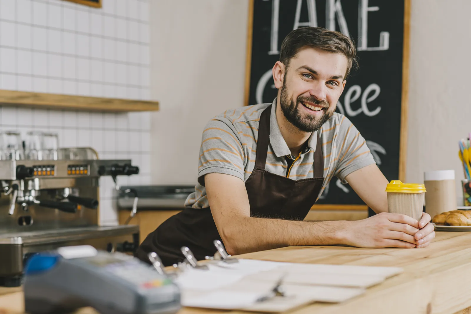 Smiling barista in an apron leaning on a café counter holding a takeaway coffee cup, with an espresso machine in the background.