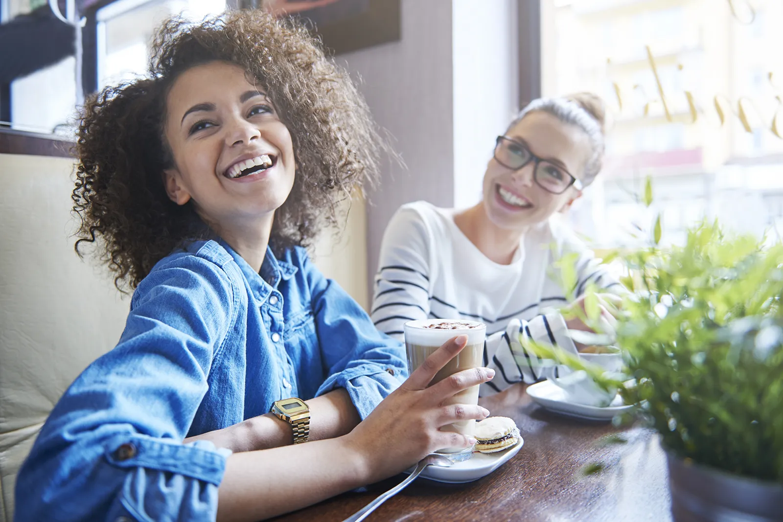 Two women sitting at a café table, smiling and laughing while enjoying coffee and pastries in a bright, cozy setting.