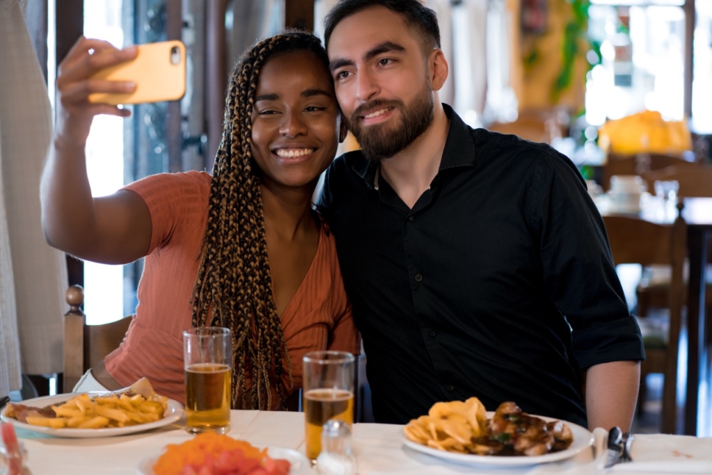 a photo of a smiling couple taking a selfie together at a restaurant table with plates of food and glasses of beer in front of them.