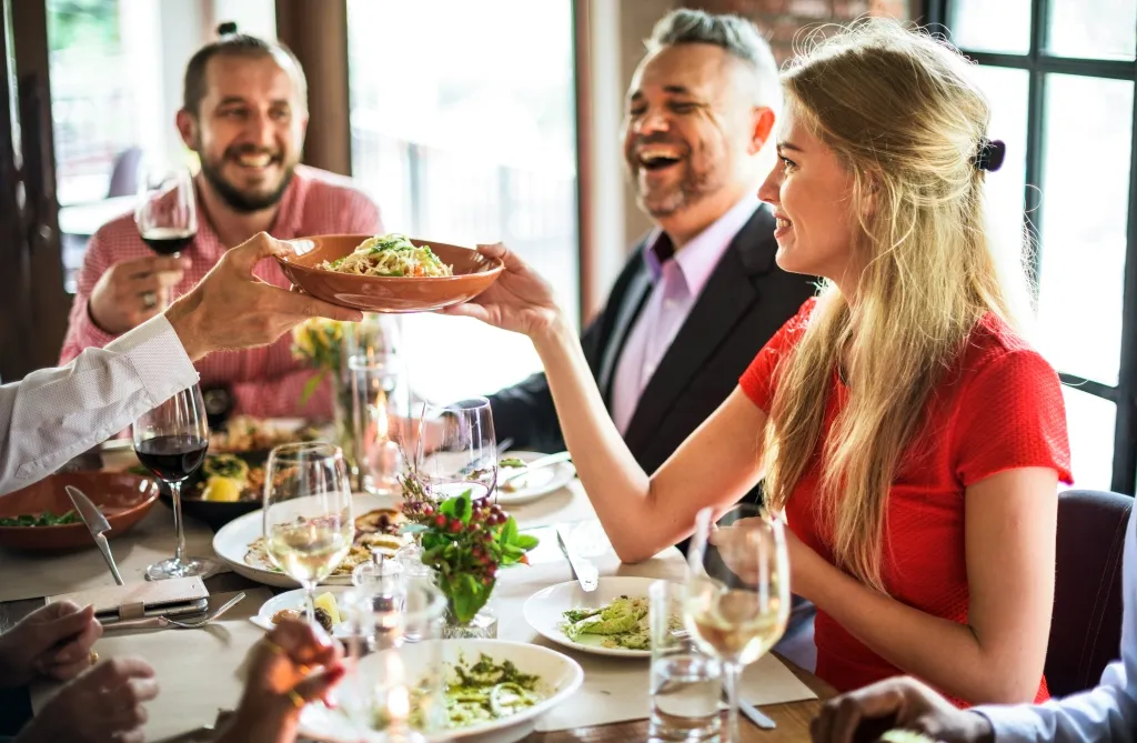 a photo of friends sharing a meal and laughing at a restaurant, with one woman in a red dress passing a bowl of pasta across the table.