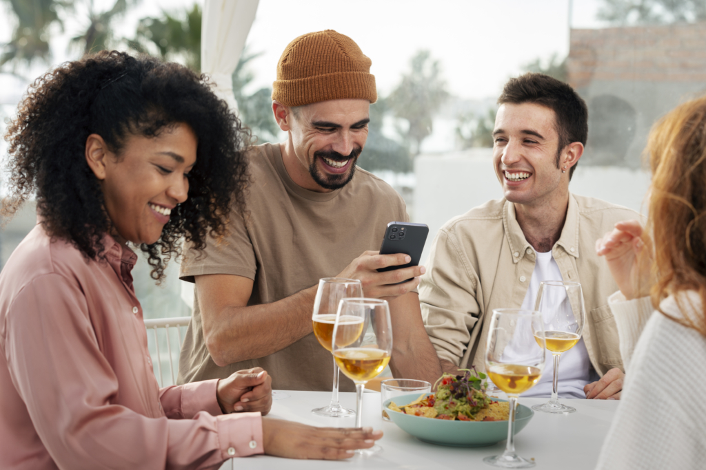 a photo of three friends sitting outdoors at a restaurant, laughing as one shows something on his phone, with drinks and nachos on the table.