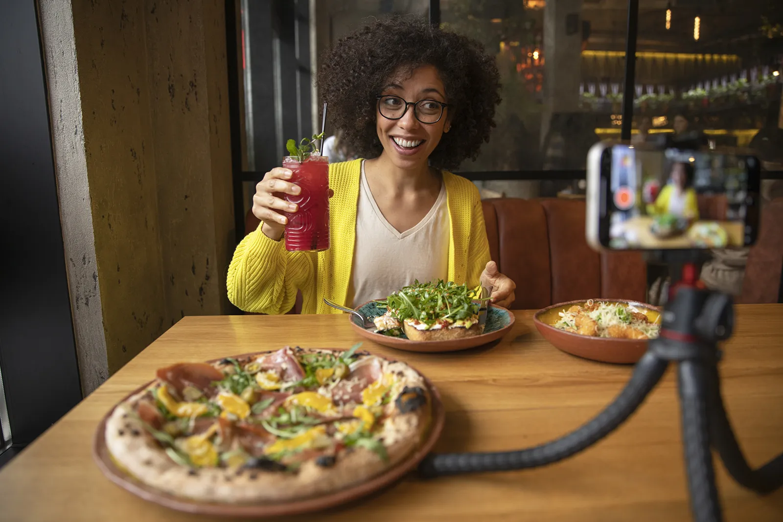 Woman smiling and holding up a red drink while filming herself with a smartphone on a tripod at a restaurant table with pizza and plated dishes.