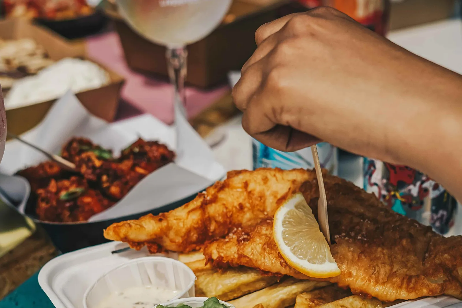 Close-up of a hand using a wooden fork to eat battered fish and chips topped with a lemon wedge, with dipping sauce and side dishes on a table.