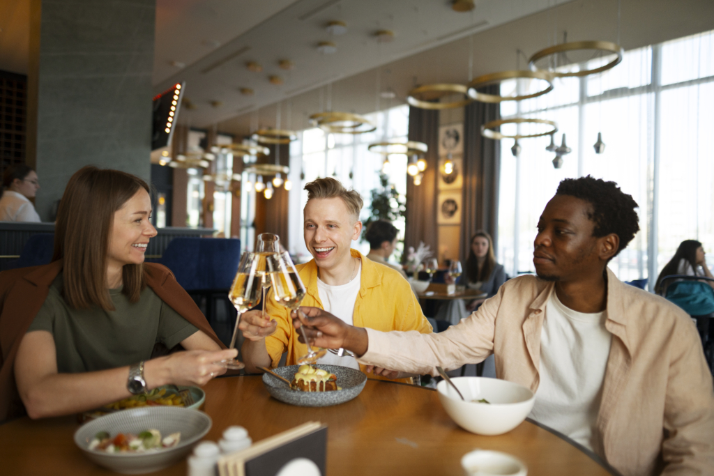 a photo of three friends clinking glasses and smiling while enjoying a meal together in a bright, modern restaurant.