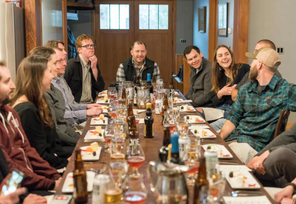a photo of a large group of people seated around a long table at a restaurant, sharing food and drinks while talking and laughing.