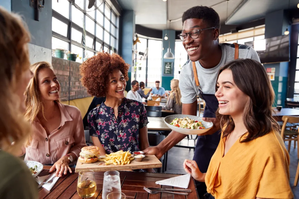 a photo of friends dining together while a smiling server delivers a dish at a bright, modern restaurant.