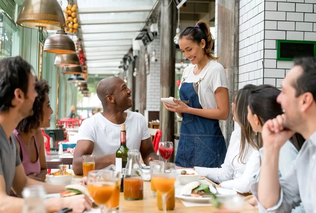 a photo of a server taking an order from a group dining at an open, sunlit restaurant patio.