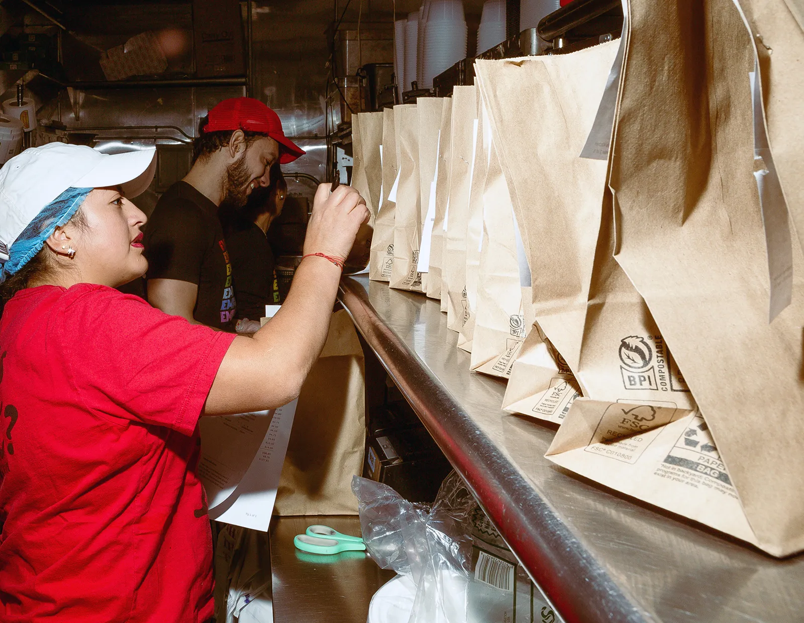 Restaurant staff organizing and labeling a row of paper takeout bags on a stainless steel counter in a busy kitchen.