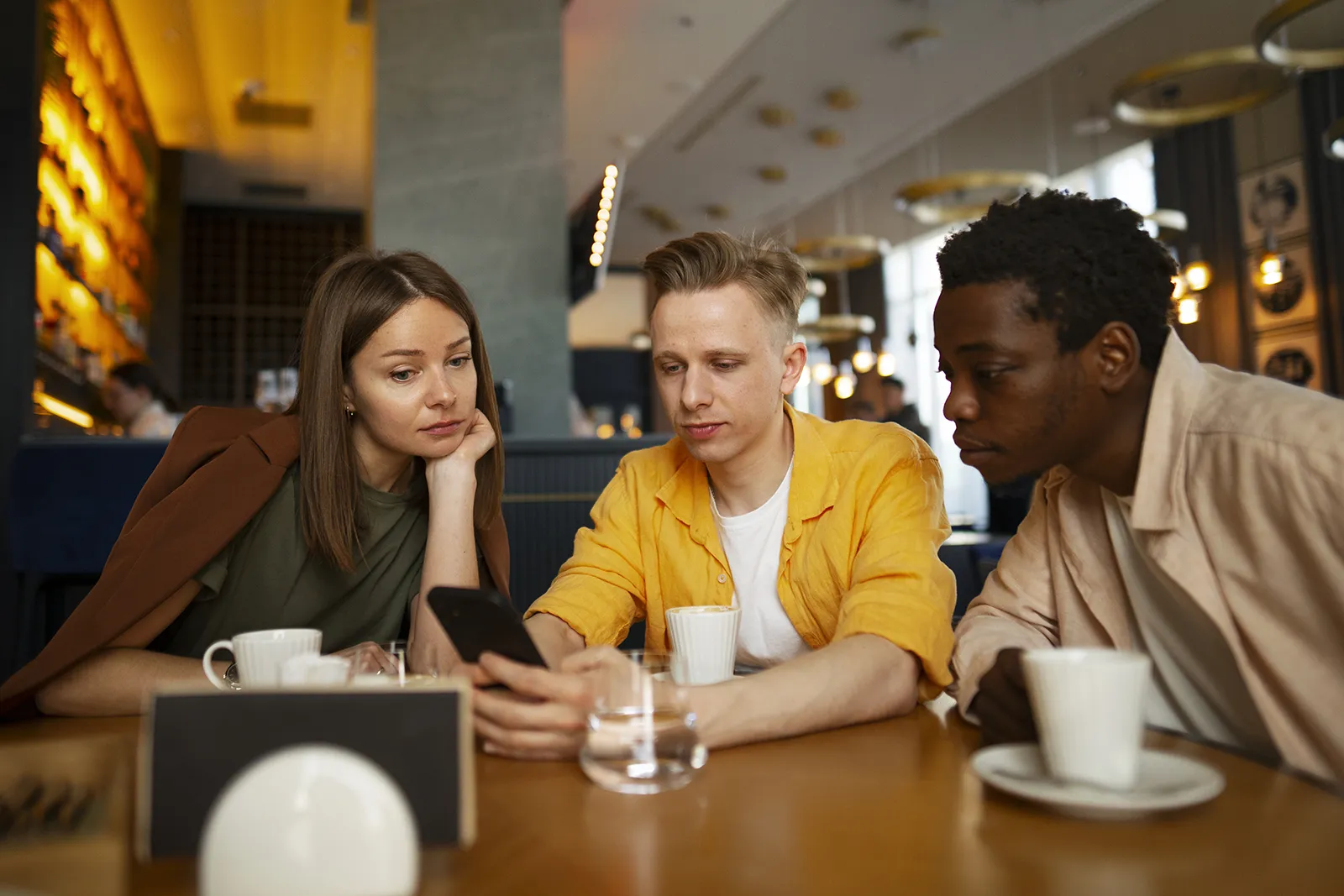 Three friends sitting at a café table, looking at a smartphone together while having coffee.
