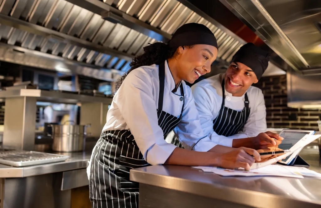a photo of two restaurant staff members reviewing orders together on a tablet in a commercial kitchen, showing teamwork and streamlined online ordering during service.