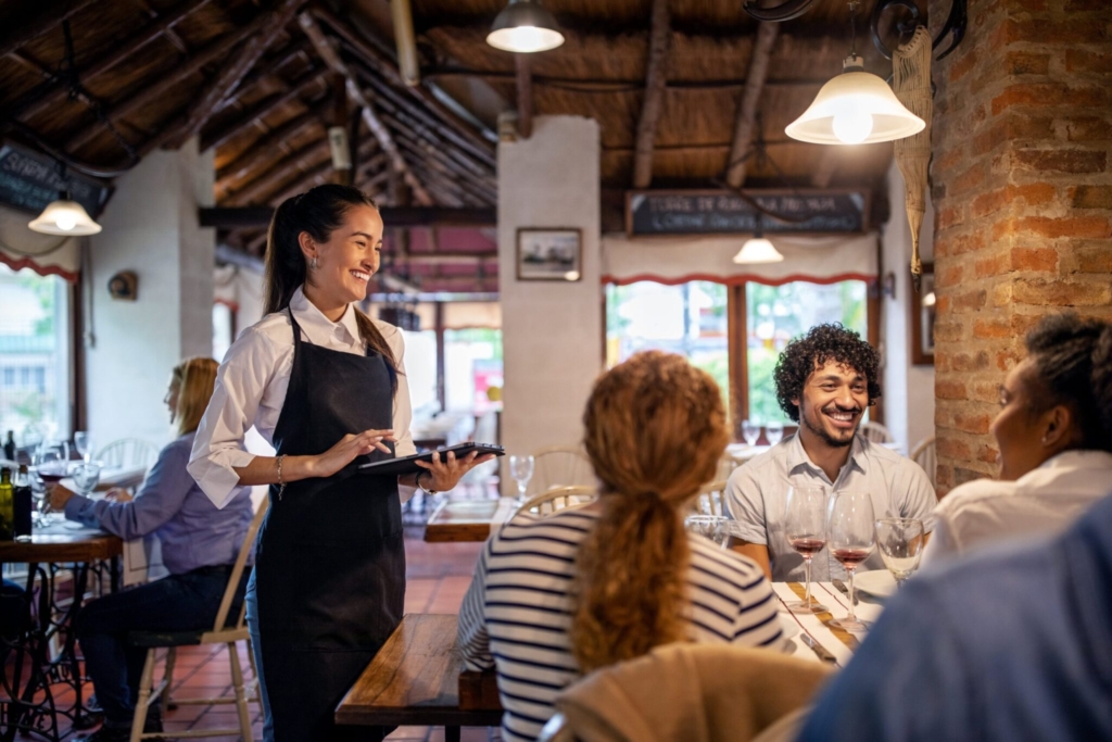 A restaurant server takes an order on a tablet while guests smile at their table, illustrating smoother front-of-house operations supported by unified order aggregation.