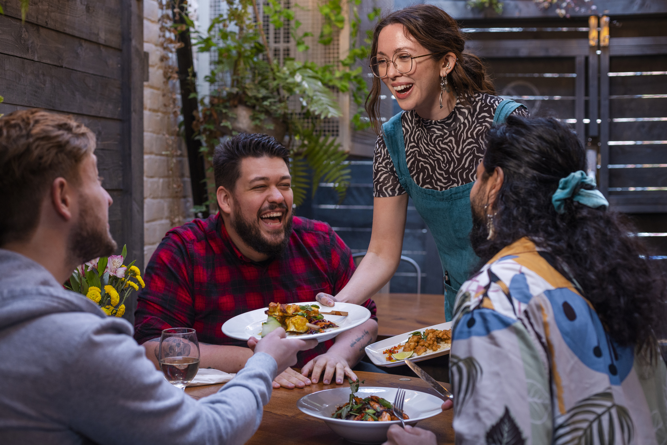 A server placing appetizer plates on a table for a group of three happy diners laughing at an outdoor restaurant table.
