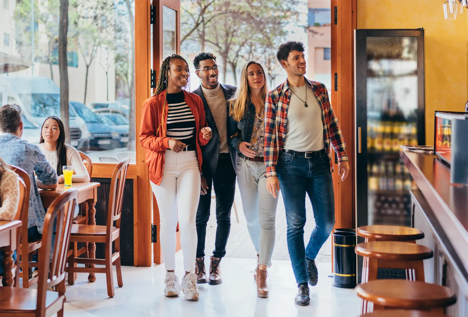 Group of four friends walking into a bright, modern restaurant, smiling and chatting as other diners sit at tables nearby.
