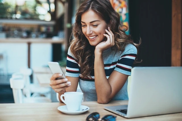 A diner sitting at a cafe table with a coffee and laptop, smiling as she looks at her smartphone. Reflects how guests prefer fast, frictionless ordering on their phones and supports the blog’s message about mobile apps improving the customer experience.