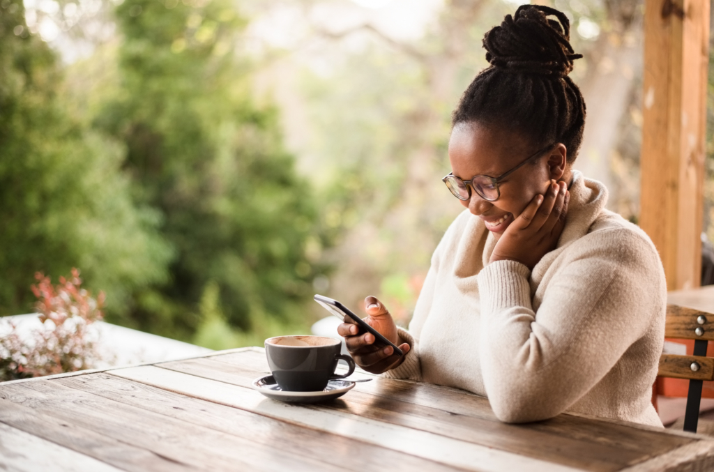A guest sits outdoors smiling at her phone while enjoying a coffee, showing how mobile ordering apps meet modern diner expectations for fast, convenient ordering