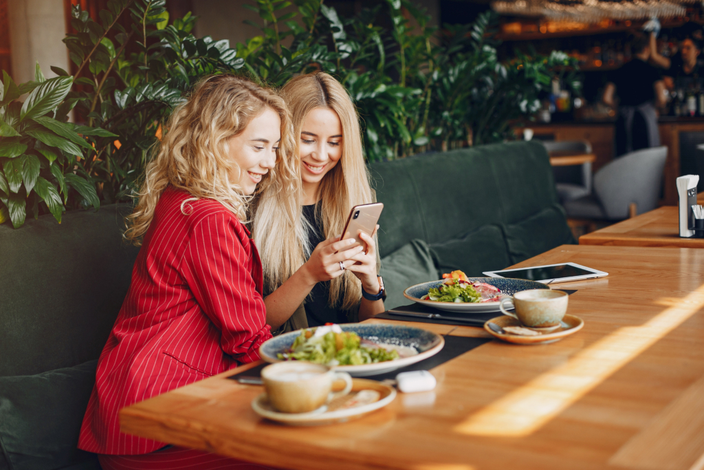 a photo of two women smiling and looking at a phone while eating salads at a restaurant table.