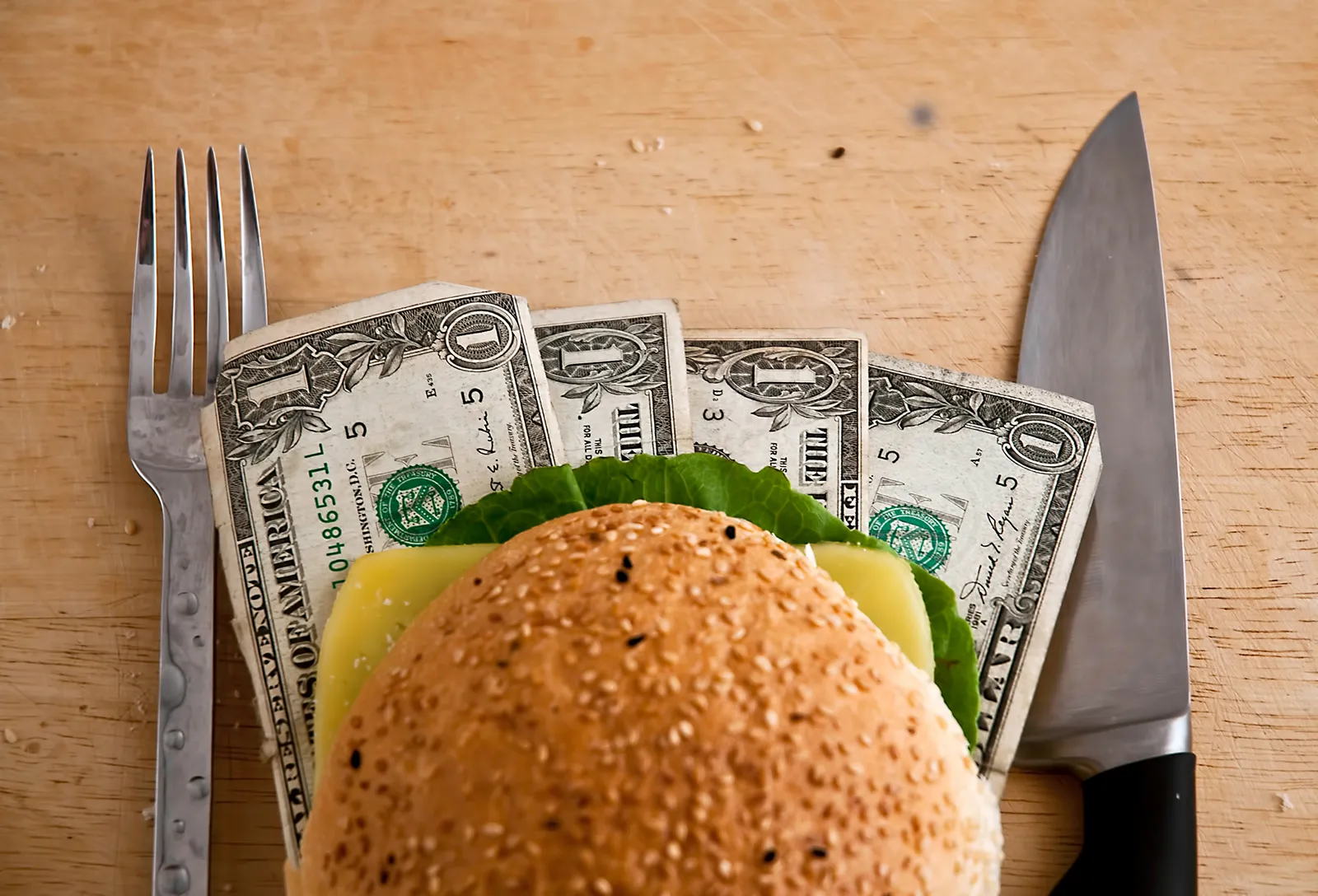Burger on a wooden cutting board with dollar bills underneath, placed between a fork and a knife, symbolizing the cost of food or dining.