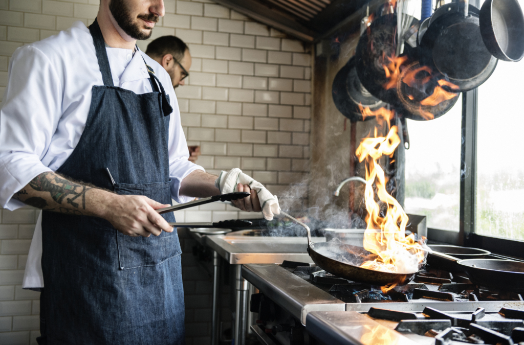a photo of a chef working over a stove with a large open flame in a busy restaurant kitchen.