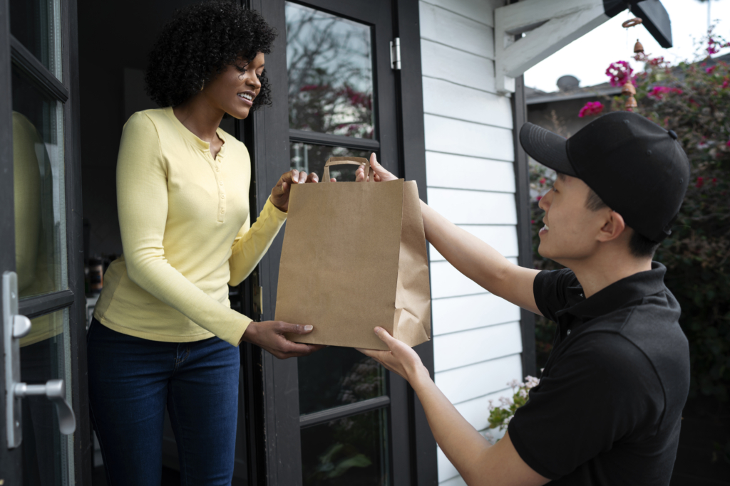a photo of a delivery driver handing a brown paper takeout bag to a woman at her front door.