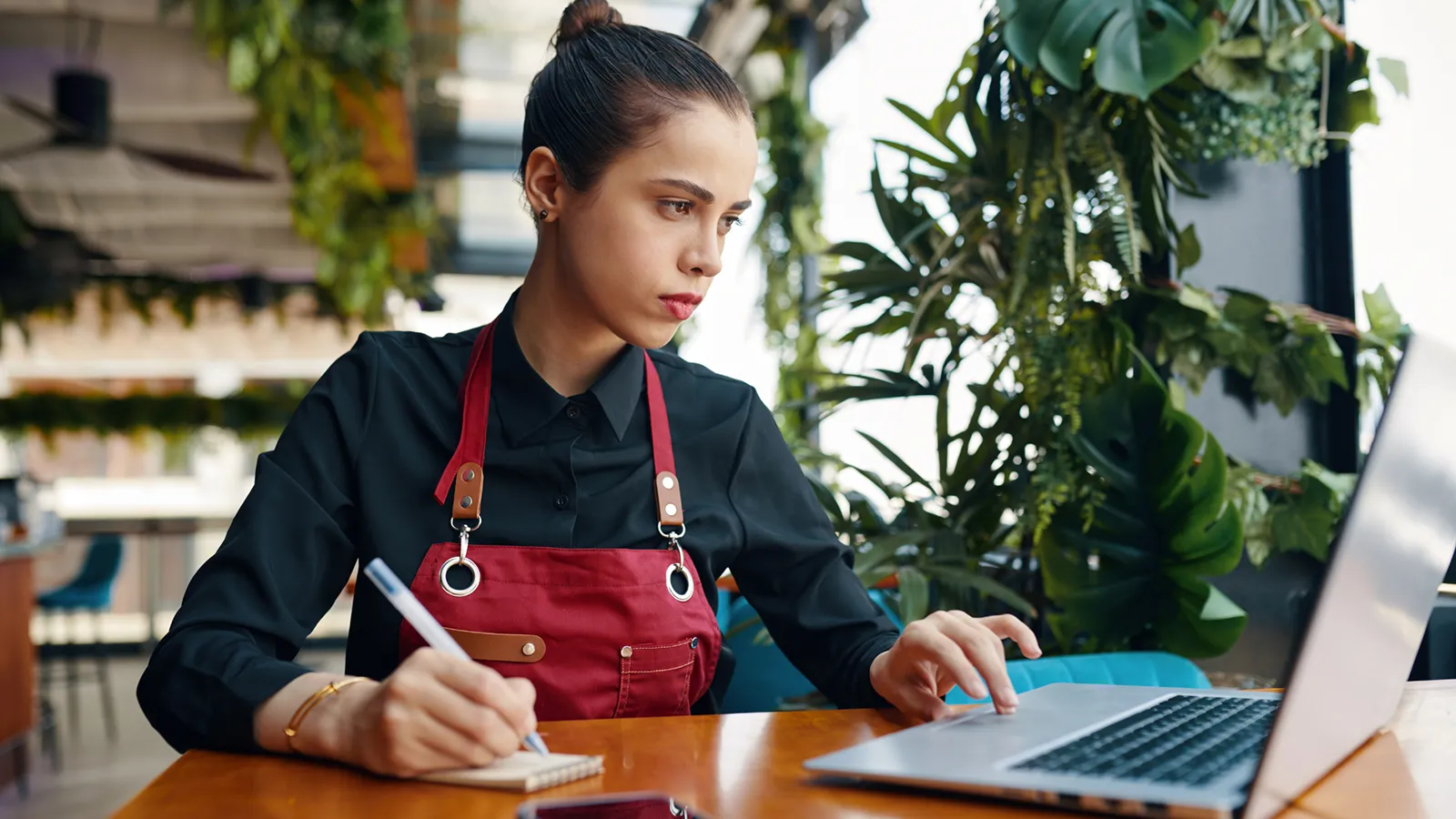 Restaurant worker wearing an apron using a laptop and writing in a notebook at a table surrounded by indoor plants.