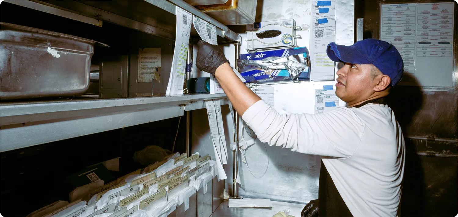 Restaurant kitchen worker pulling a printed order ticket from a rack in a busy prep area, with shelves of containers and order slips in the background.