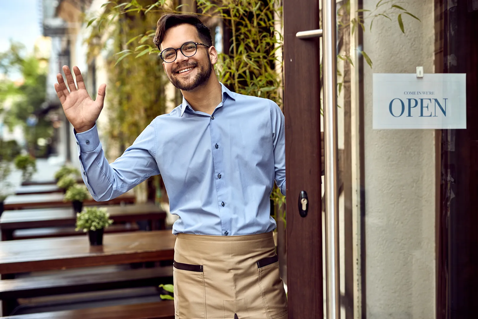 Restaurant owner or staff member standing in a doorway, smiling and waving to customers, with an ‘Open’ sign visible on the door and outdoor tables in the background.