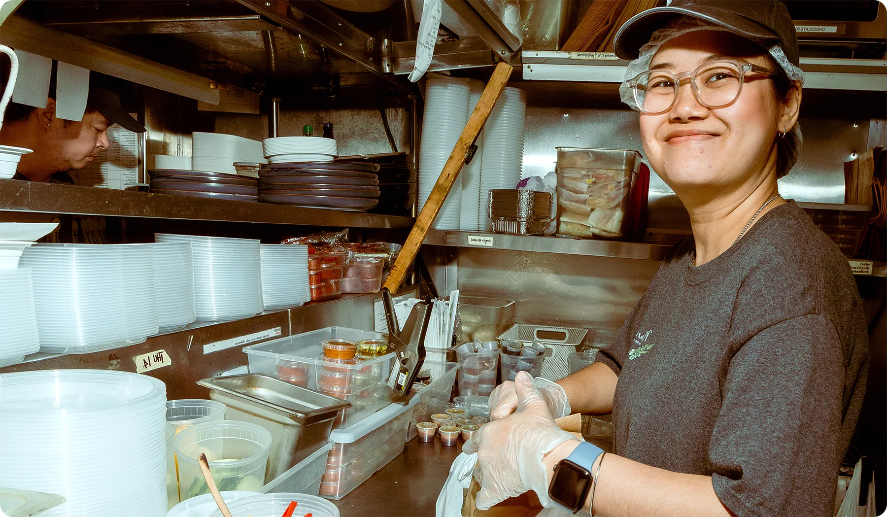 Smiling restaurant worker wearing a cap, glasses, and gloves preparing takeout items in a busy kitchen prep station with containers, sauces, and stacked plates.