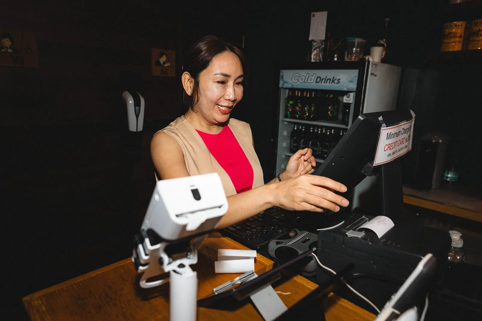 Smiling woman in a red dress using a tablet at a bar counter with a POS system and card reader, preparing to take an order.