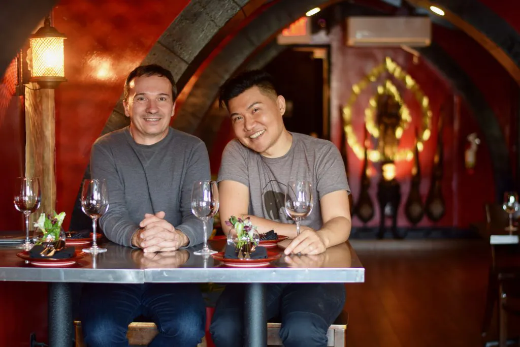 Two men sitting side by side at a restaurant table with wine glasses, smiling at the camera in a warmly lit red interior with arched ceilings.