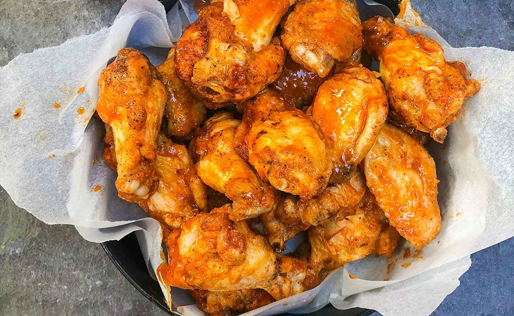Basket of crispy chicken wings coated in orange buffalo sauce, served on parchment paper.