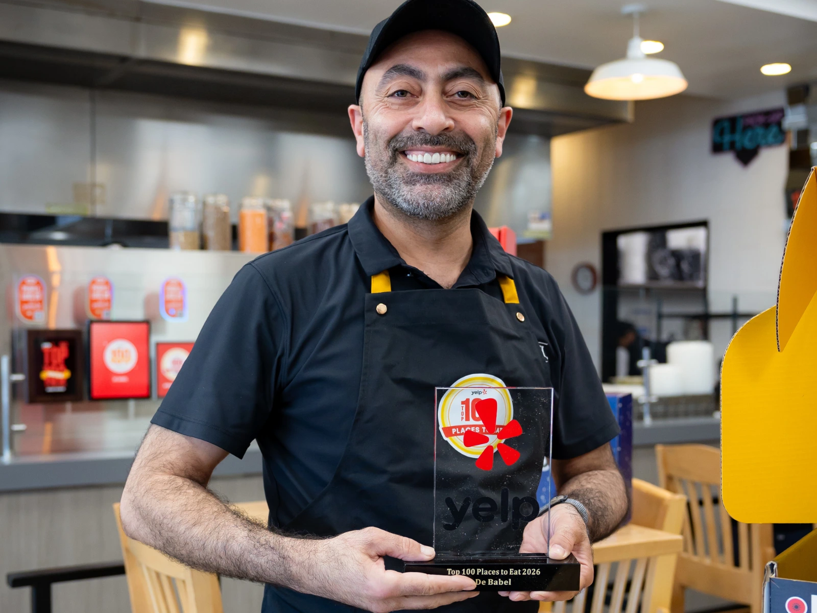 Marwan Kandeel, owner of De Babel, smiling and holding a Yelp Top 100 Places to Eat 2026 award in his restaurant