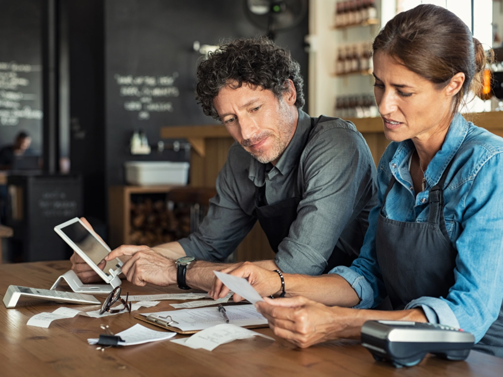 Two restaurant owners reviewing receipts and sales data together at a counter, using a tablet and calculator to track online ordering revenue.